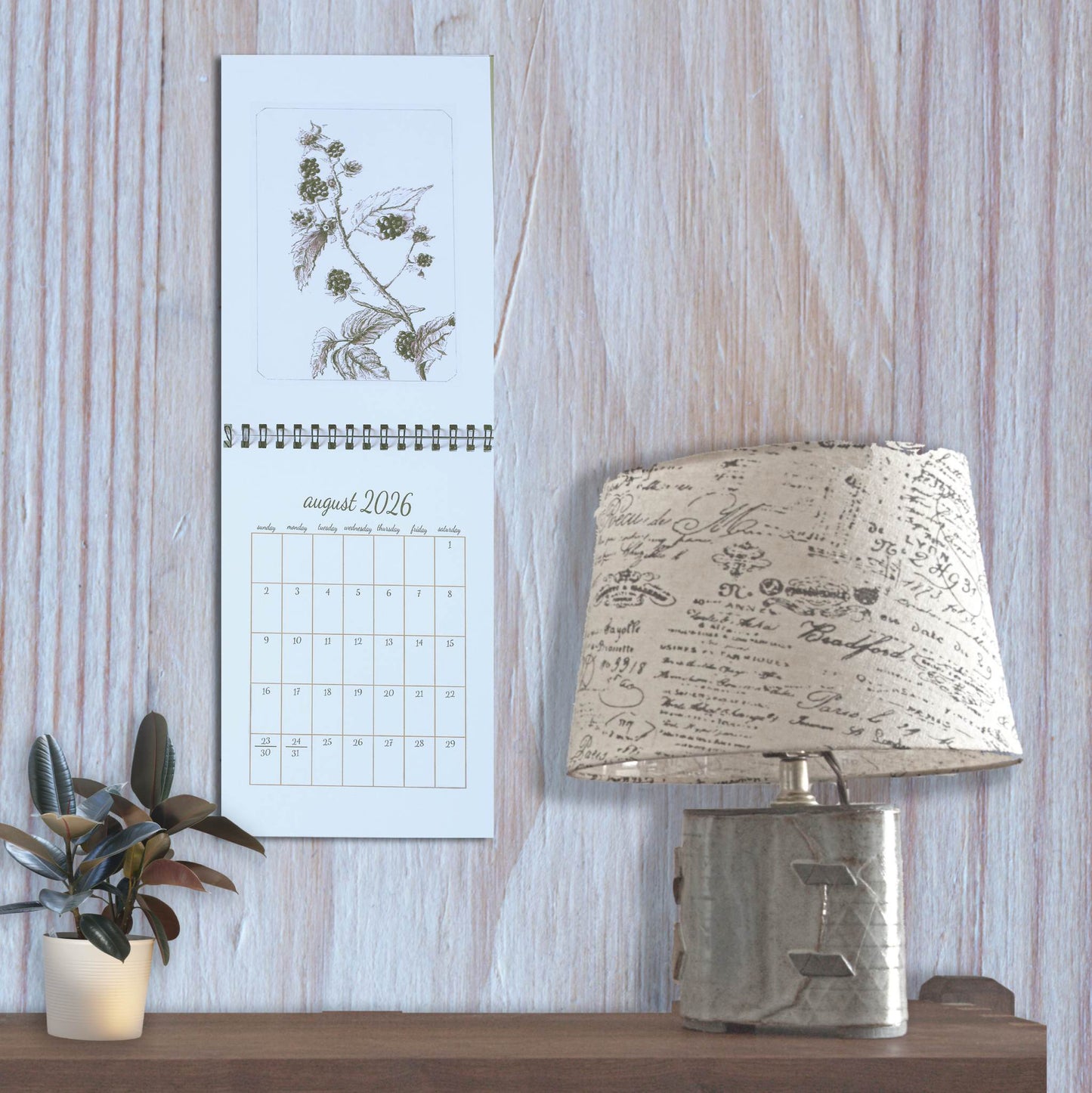 2026 inked woods wall calendar displayed on a wall, styled with a lamp on the right and a small plant on the left.