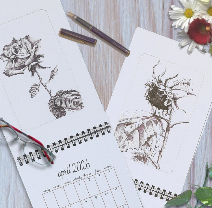 2026 art wall calendar on a wooden surface open to two months, featuring original ink and watercolor drawings of a rose and a sunflower, styled with fresh flowers, glasses, a fountain pen and some greens.