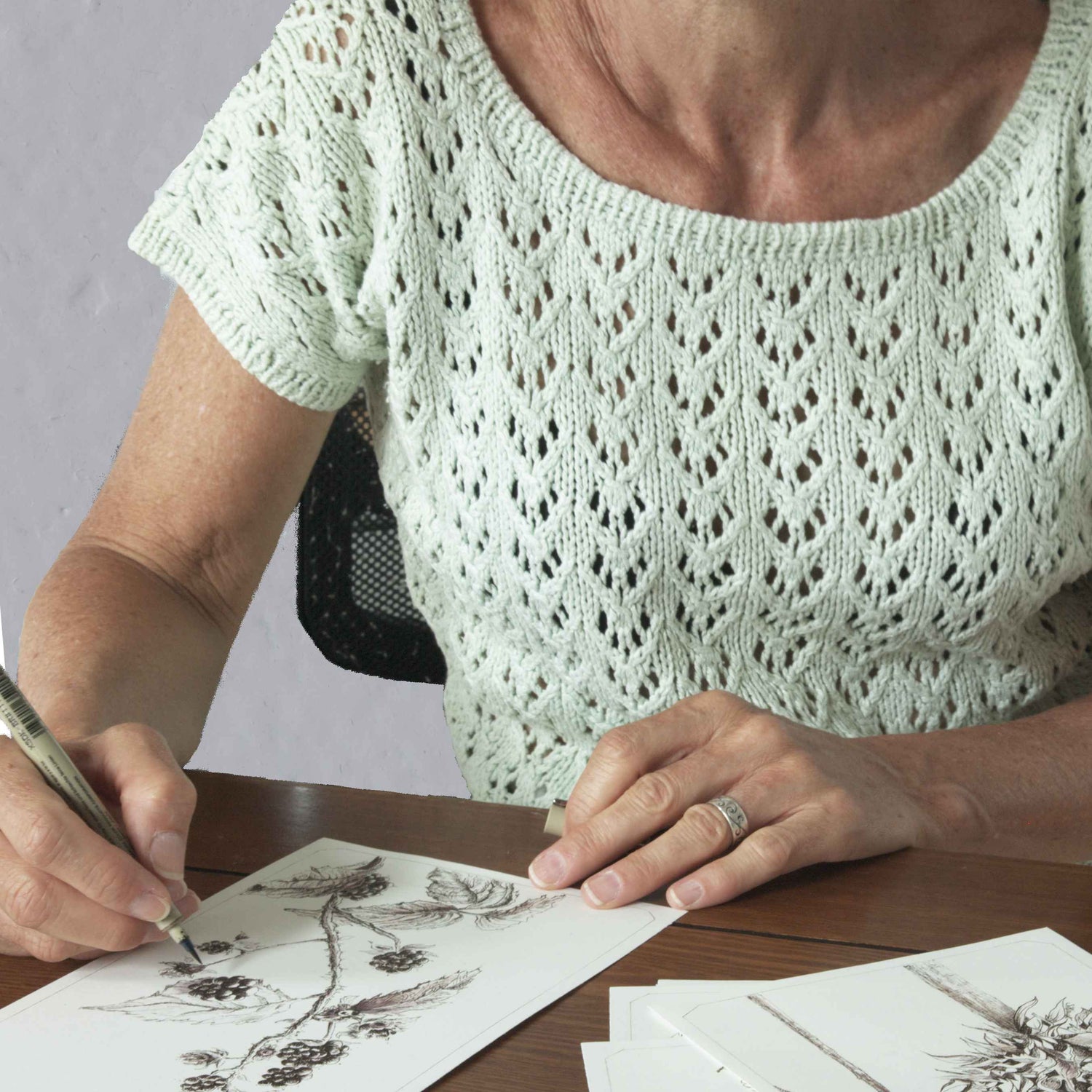 Artist in light green lace top drawing blackberries with ink pen at desk, original artwork in progress.
