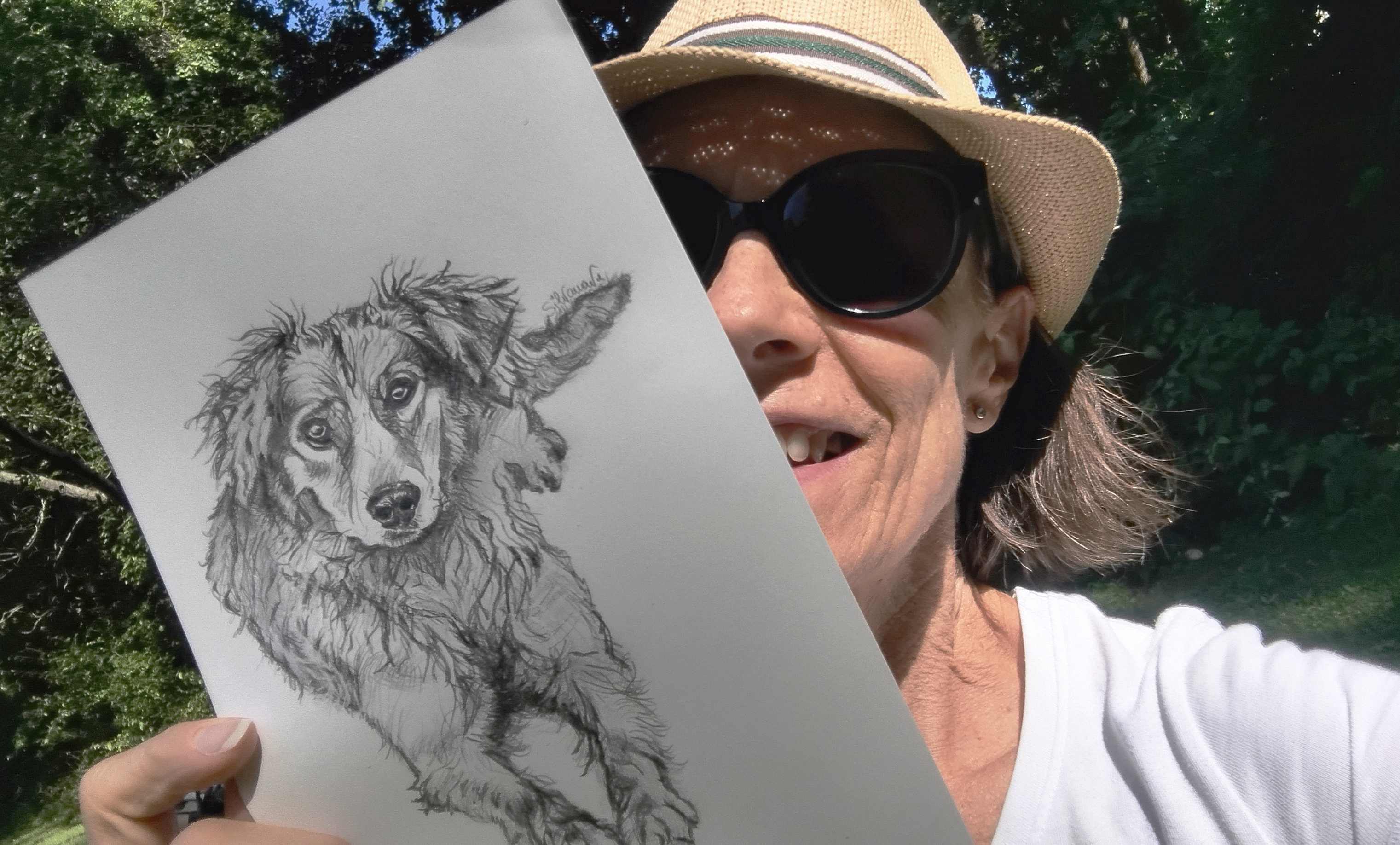 Artist outdoors wearing a panama hat and sunglasses, holding a pencil dog portrait in hands, artwork partly hiding face