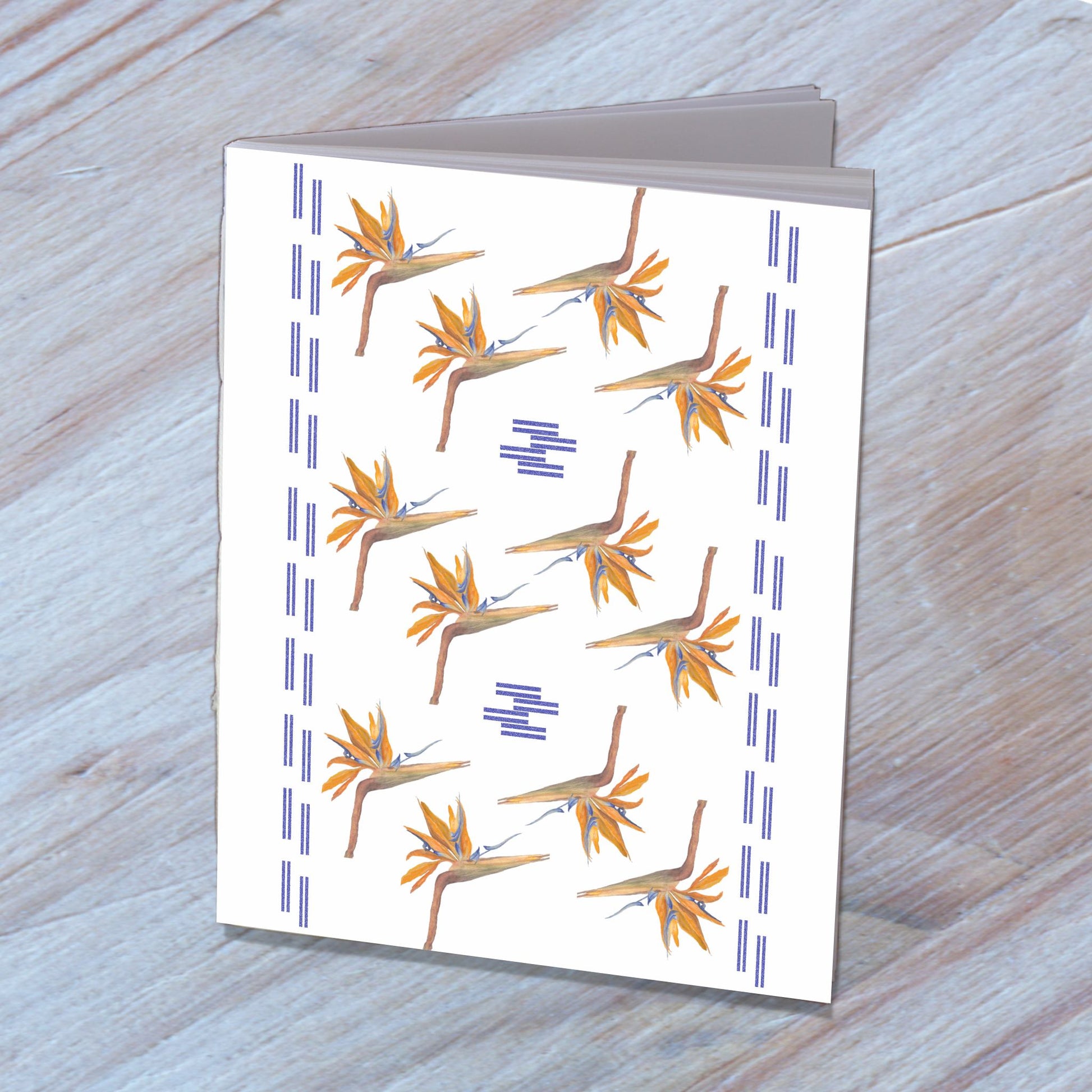 Handmade notebook with a cover designed from bird of paradise flower watercolor, standing on a wooden surface.
