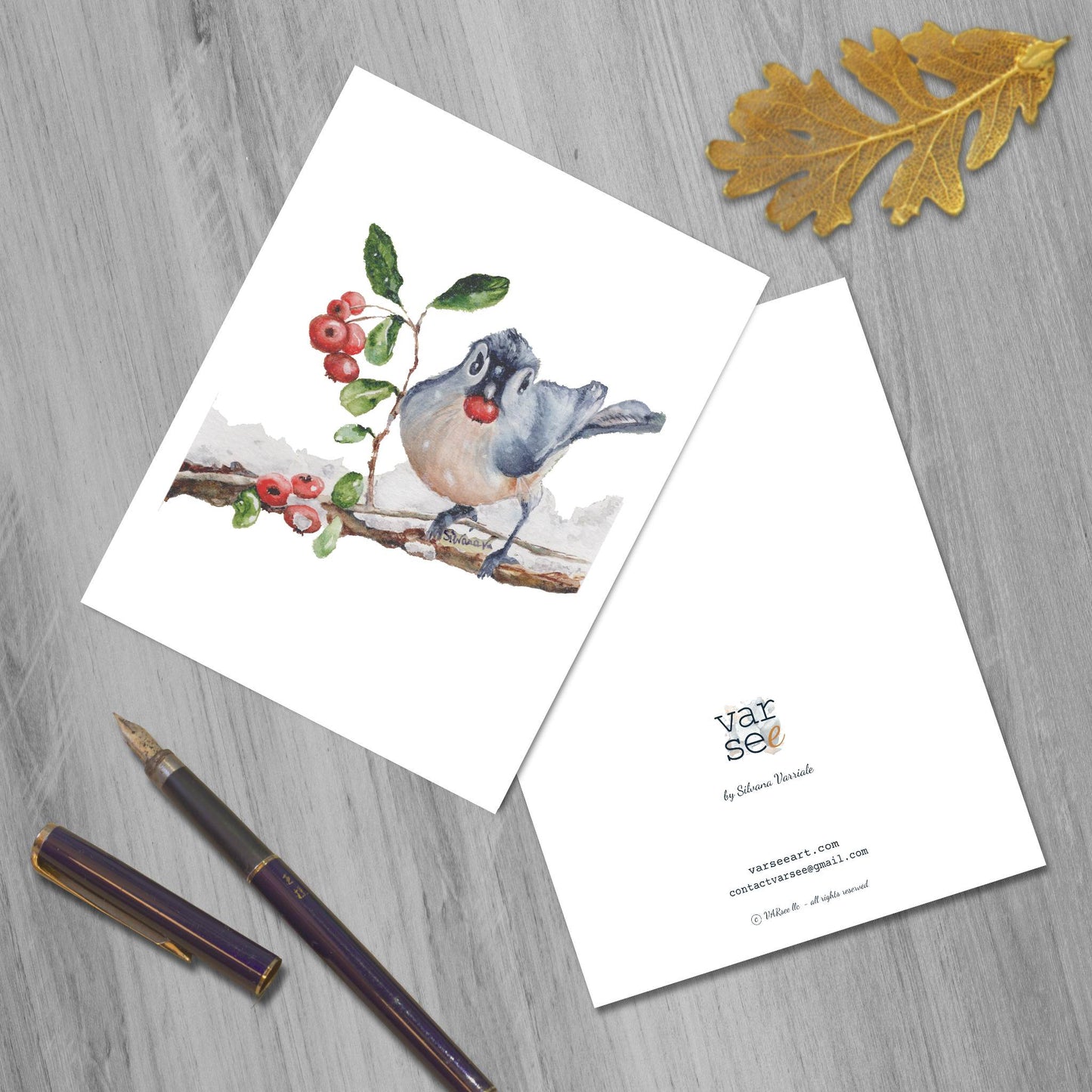 Front and back of a blue jay bird eating a berry watercolor greeting card on a flat surface, a golden leaf and a fountain pen beside.