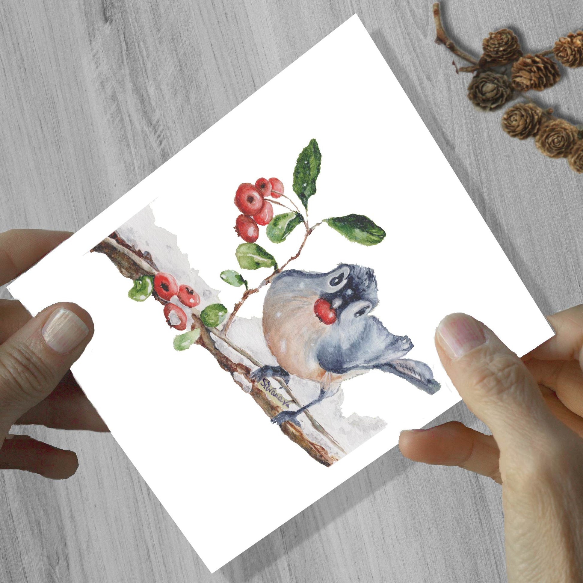 Hands holding a blue jay bird eating a berry watercolor greeting card, shown on a wooden surface.