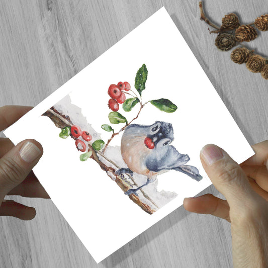Hands holding a blue jay bird eating a berry watercolor greeting card, shown on a wooden surface.