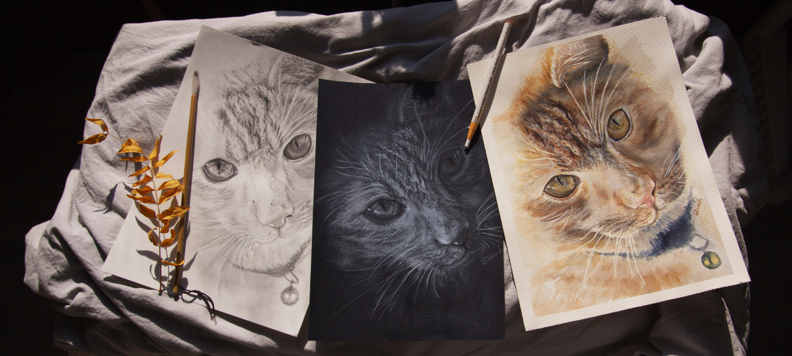 Three original portraits of the same red tabby cat, showcasing a consistent likeness in watercolor, graphite pencil, and white pencil on black paper. The artwork by Silvana Varriale of VARsee is arranged on crumpled fabric in natural sunlight with a paintbrush, a pencil, and a yellow leaf branch.