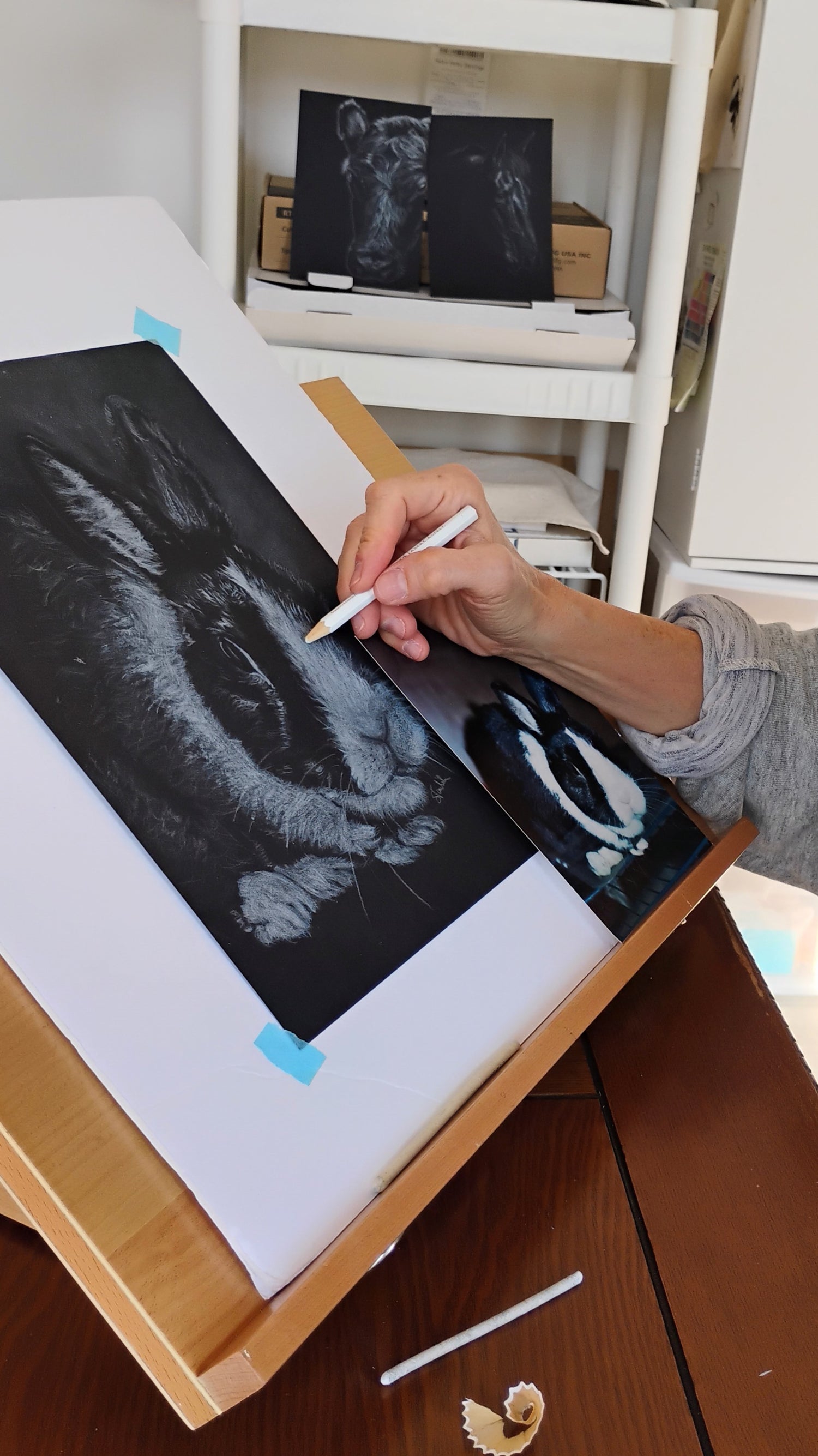 Close-up of artist Silvana Varriale of VARsee Art creating a custom rabbit memorial portrait on a wooden easel. The artist's hand uses a white pencil on black paper to draw an intricate original artwork, referencing a nearby photograph of a black and white bunny to capture the exact likeness and honor the lost pet.