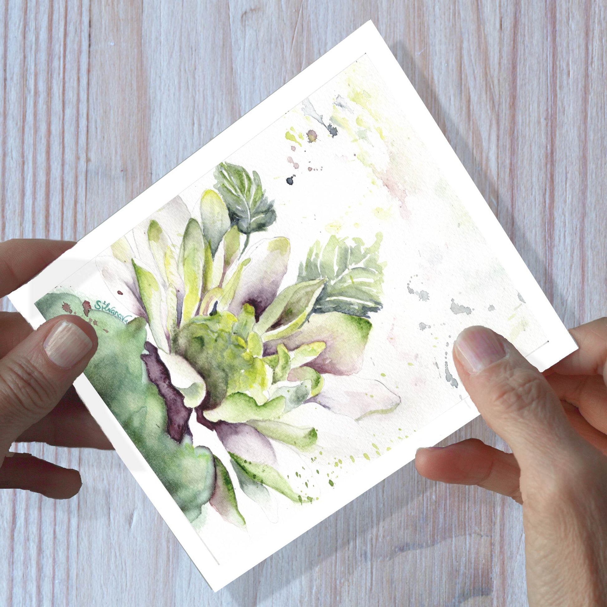 Hands holding  art of a dahlia watercolor greeting card, shown on a wooden surface.