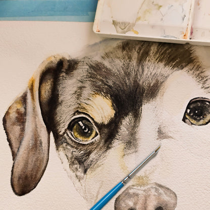 A close-up of a fine-tipped paintbrush laying across dog's portrait in watercolor. This image highlights the traditional, hand-painted nature of the varsee service, shown alongside a working watercolor palette.
