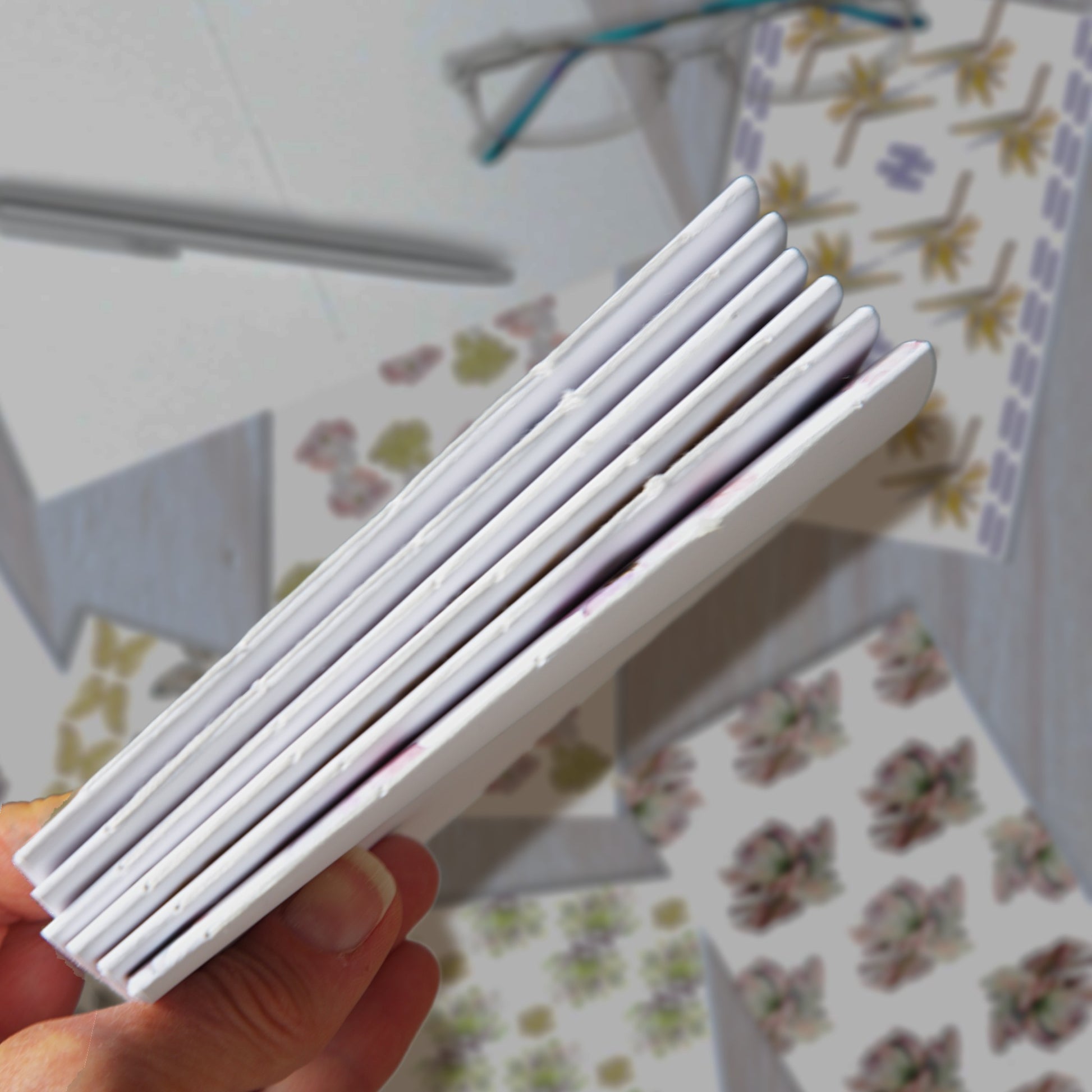 Hand holding six closed notebooks tightly together, showing the stitched bindings, with blurred notebook designs in the background.