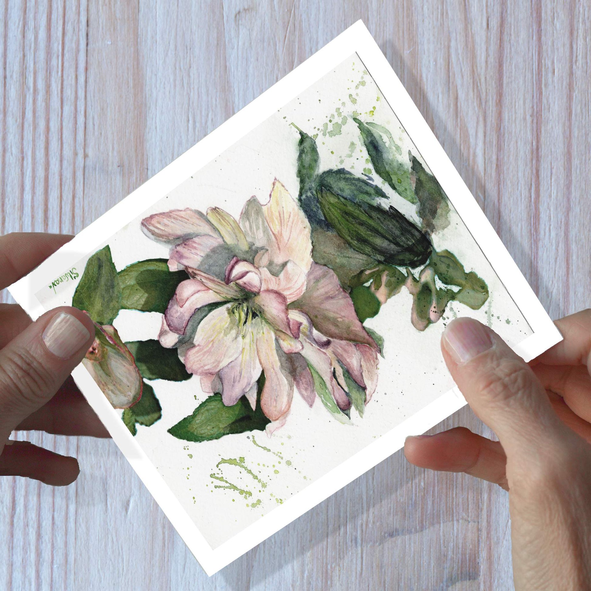 Hands holding watercolor greeting card art of a hellebore flower, shown on a wooden surface.