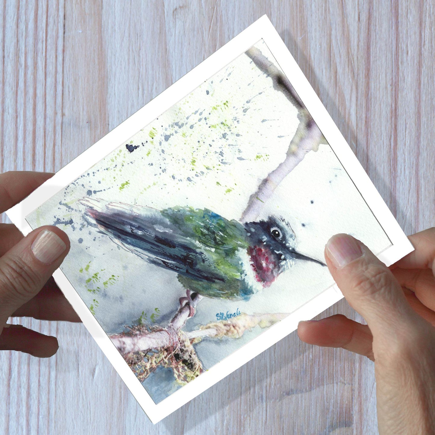 Hands holding a hummingbird watercolor greeting card, shown on a wooden surface.