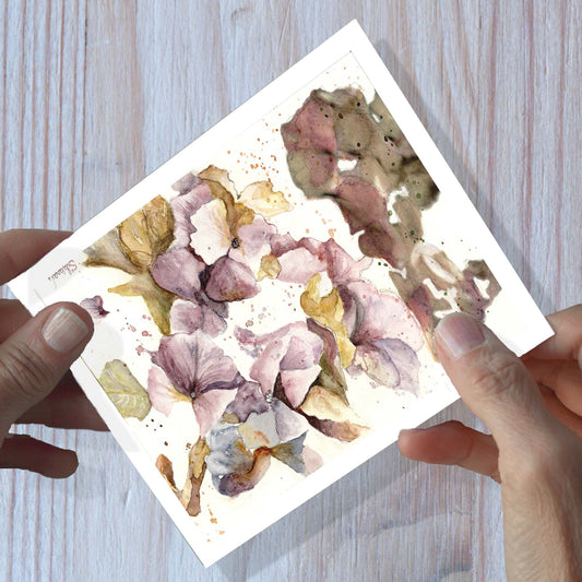 Hands holding an hydrangea watercolor greeting card, shown on a wooden surface.