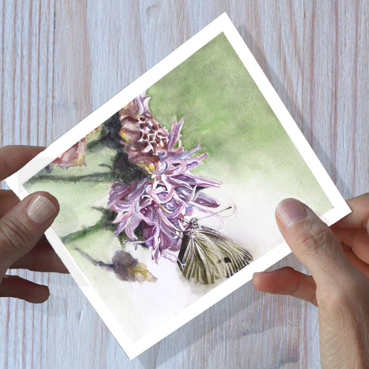 Hands holding a knapweed watercolor greeting card, shown on a wooden surface.