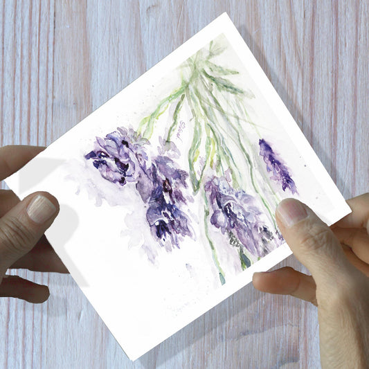 Hands holding a lavender sprig watercolor greeting card, shown on a wooden surface.