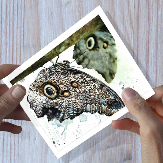 Hands holding owl eye butterfly watercolor greeting card art, shown on a wooden surface