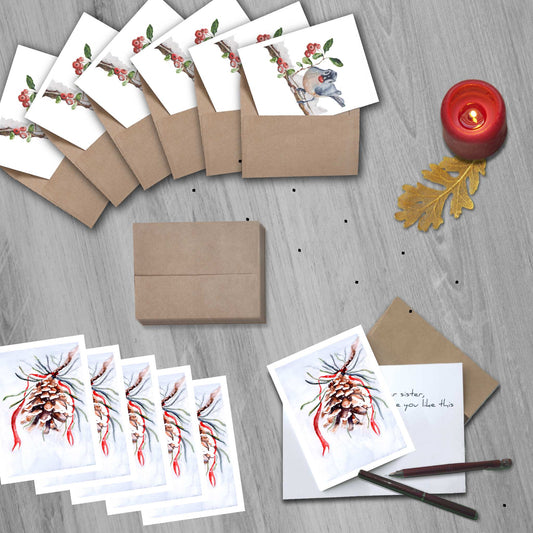 Set of 12 watercolor greeting cards (6 pinecone, 6 blue jay bird), one open on the table with a partially written message and a fountain pen on it.