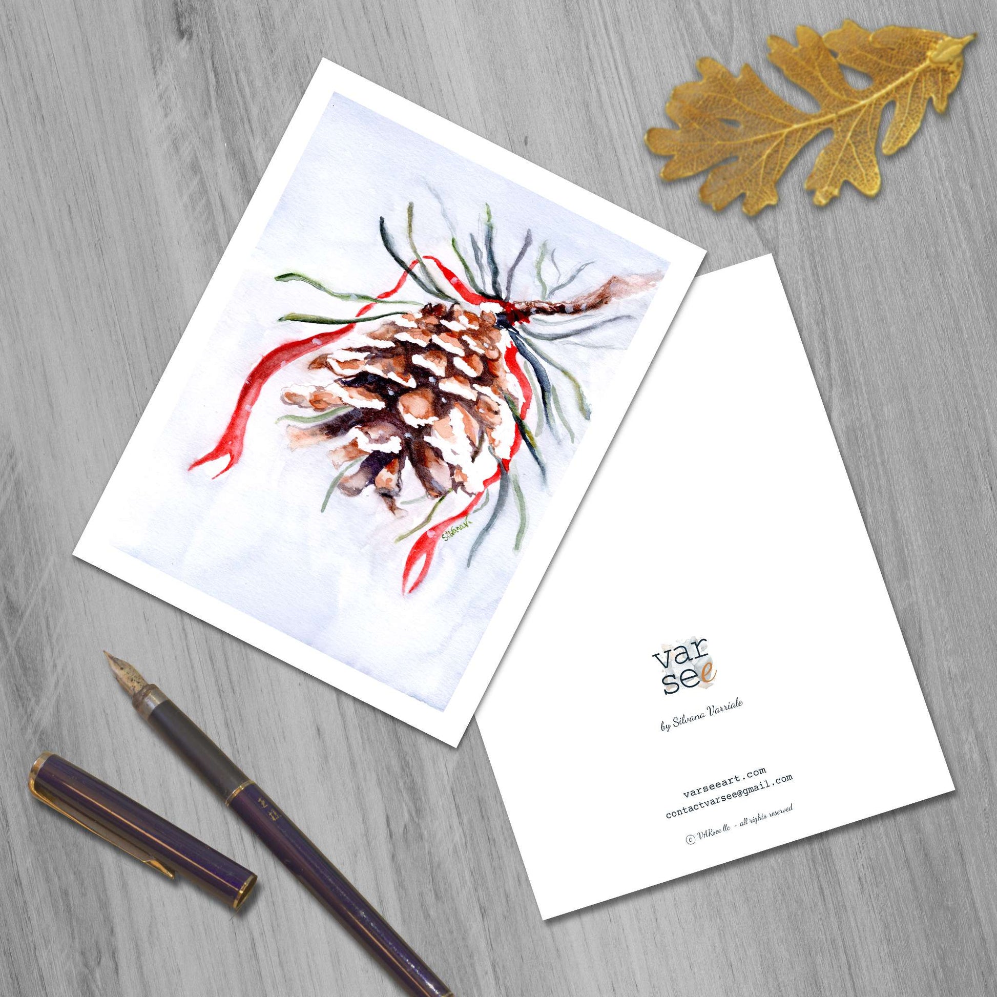 Front and back of a pinecone watercolor greeting card on a flat surface, a golden leaf and a fountain pen beside.