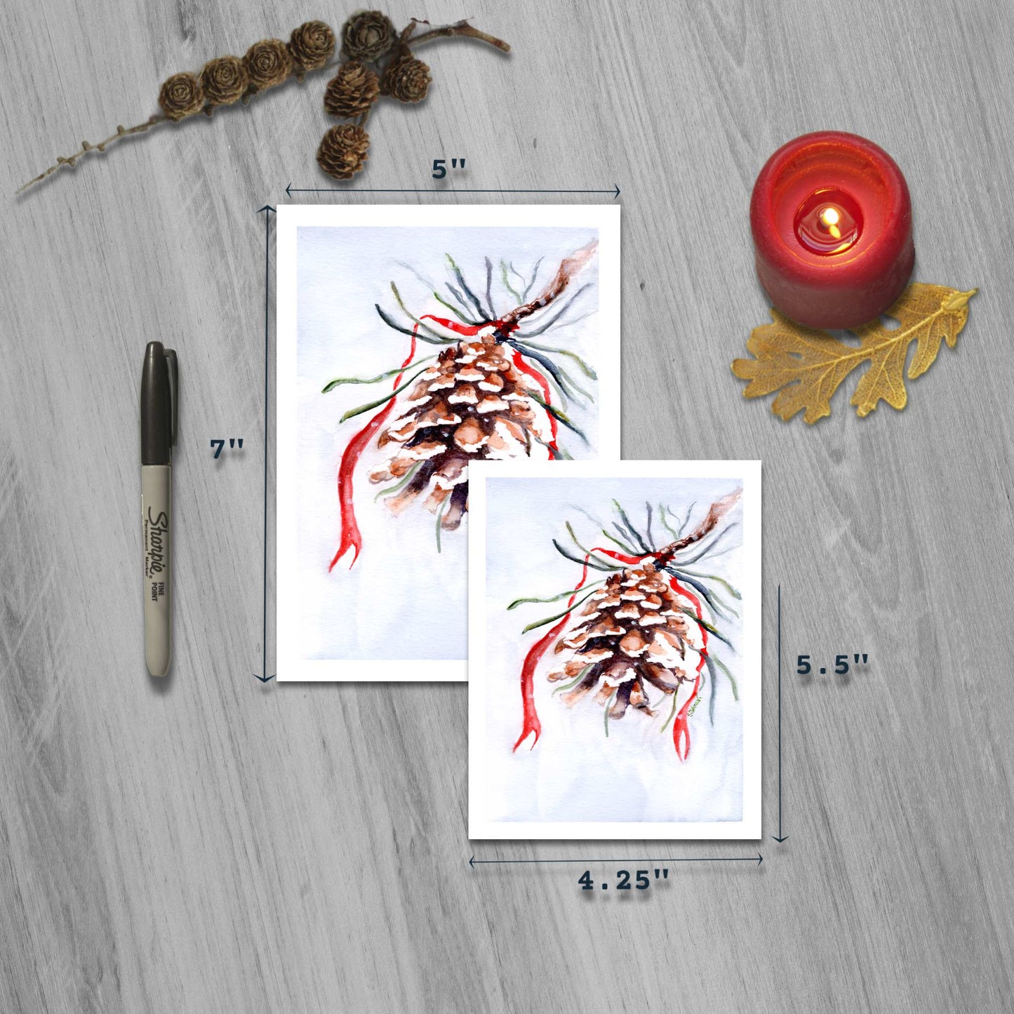 Pinecone and red ribbon watercolor greeting cards in small and large sizes with marker for scale.