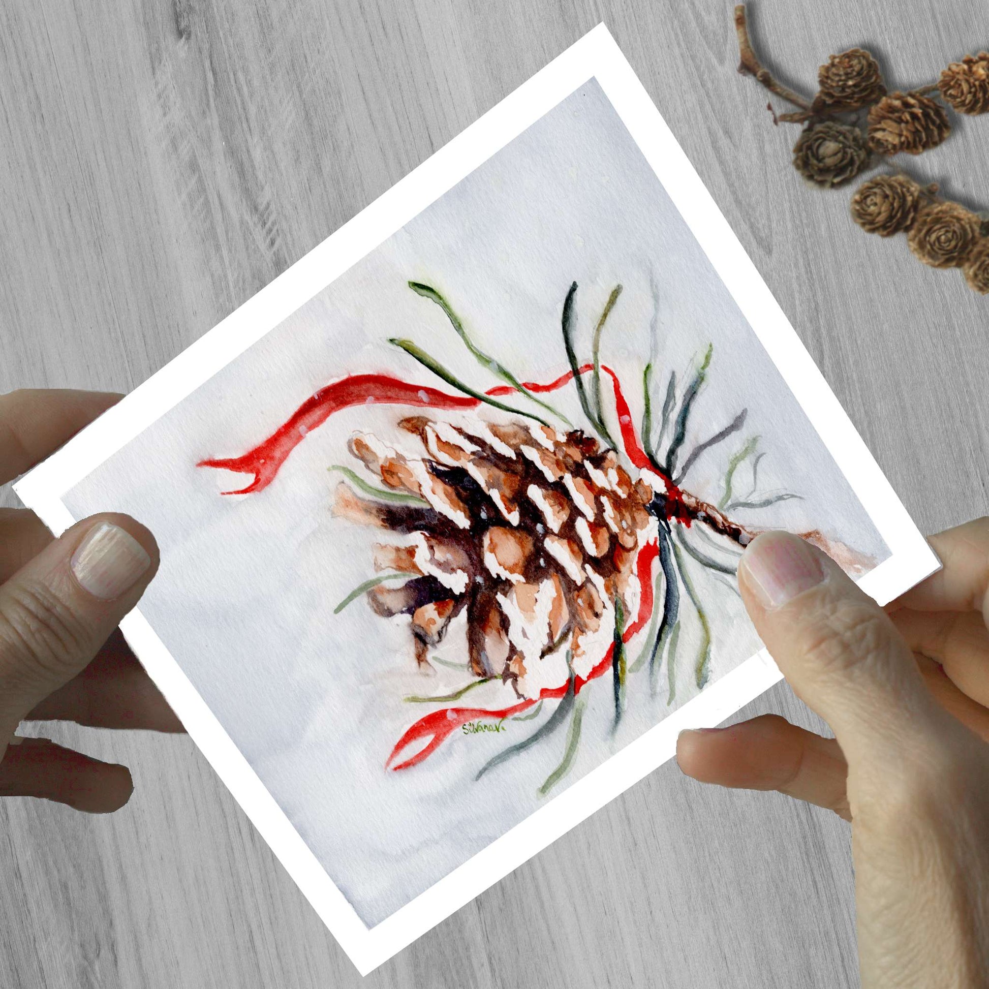 Hands holding a watercolor greeting card of a pinecone decorated with a red ribbon, shown on a wooden surface.