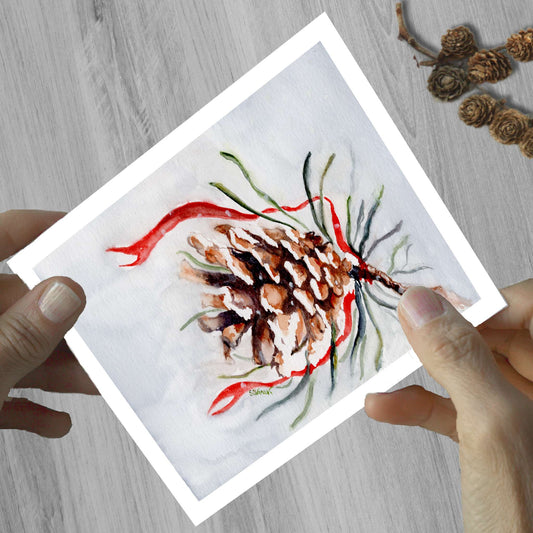 Hands holding a watercolor greeting card of a pinecone decorated with a red ribbon, shown on a wooden surface.