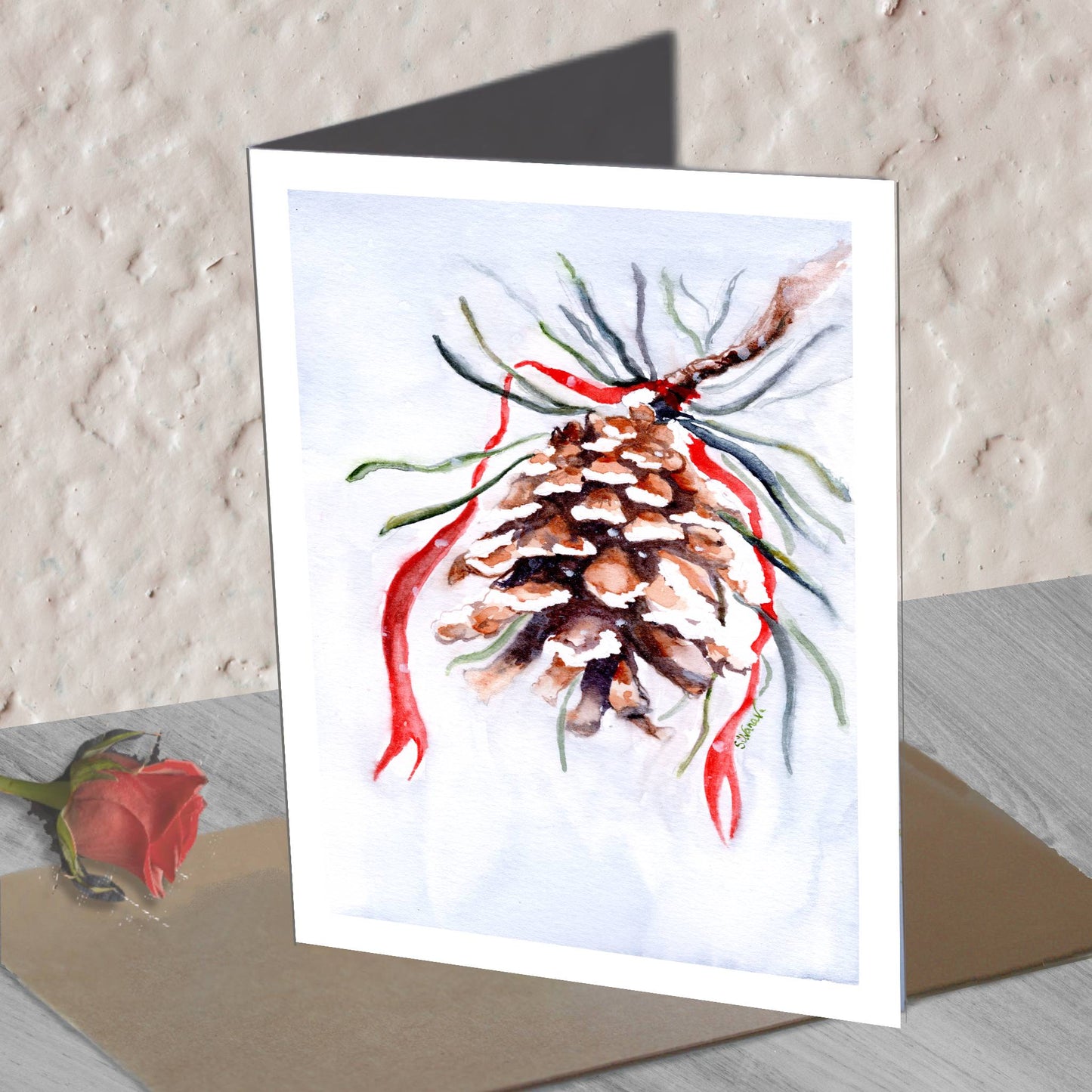 Pinecone watercolor greeting card with red ribbon standing on an envelope on a table, with a real pine branch beside.