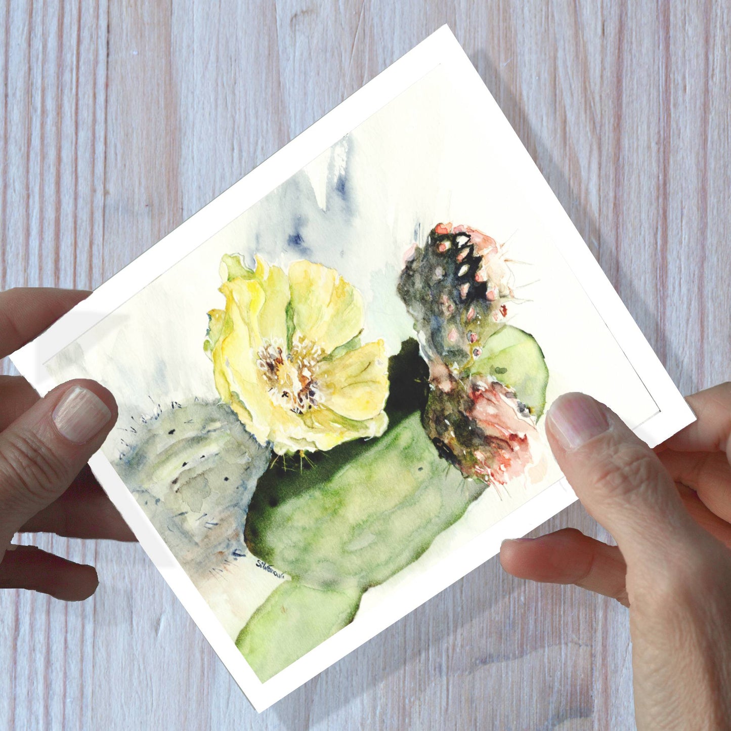 Hands holding a prickly pear watercolor greeting card, shown on a wooden surface.