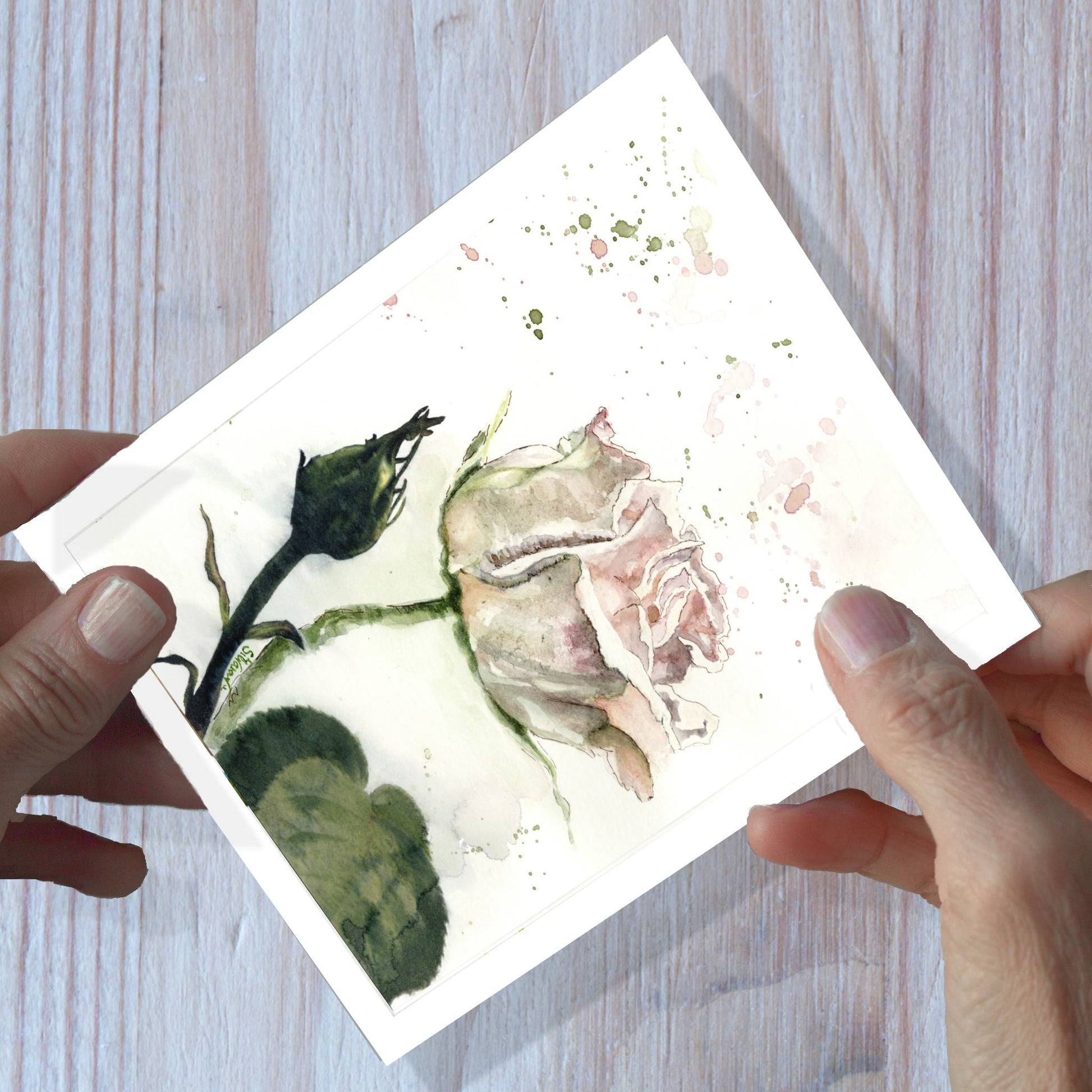 Hands holding a pink rose watercolor greeting card, shown on a wooden surface.