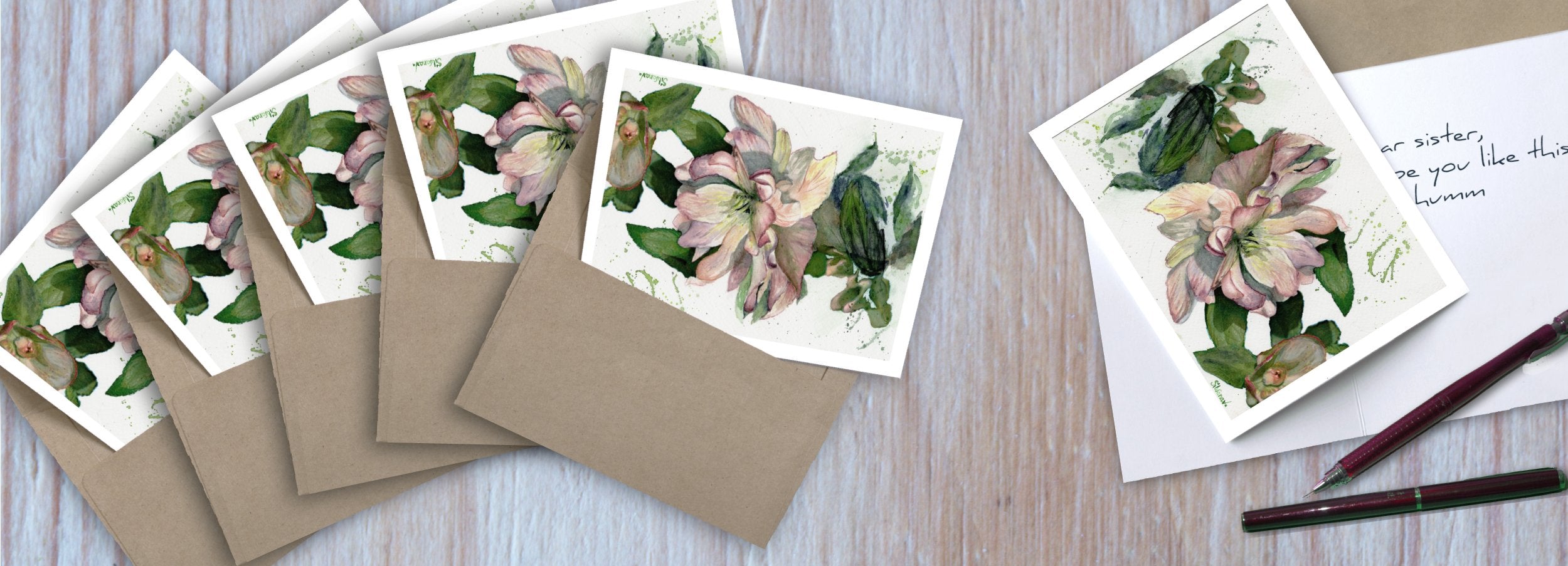Set of 5 hellebore watercolor greeting cards with envelopes, plus a sixth card open with partial message and fountain pen on wooden surface.