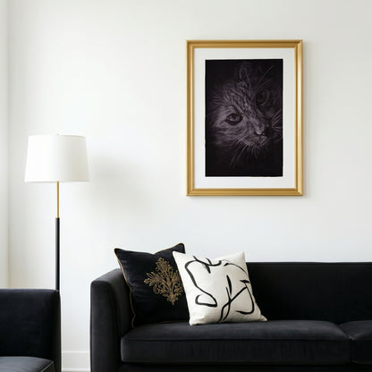 A vertical print of a Silvana Varriale original cat drawing in white pencil on white paper, professionally displayed in a minimal golden frame. The artwork is staged on a clean white wall above a sophisticated black velvet couch, fitting a modern black, white, and gold interior palette.