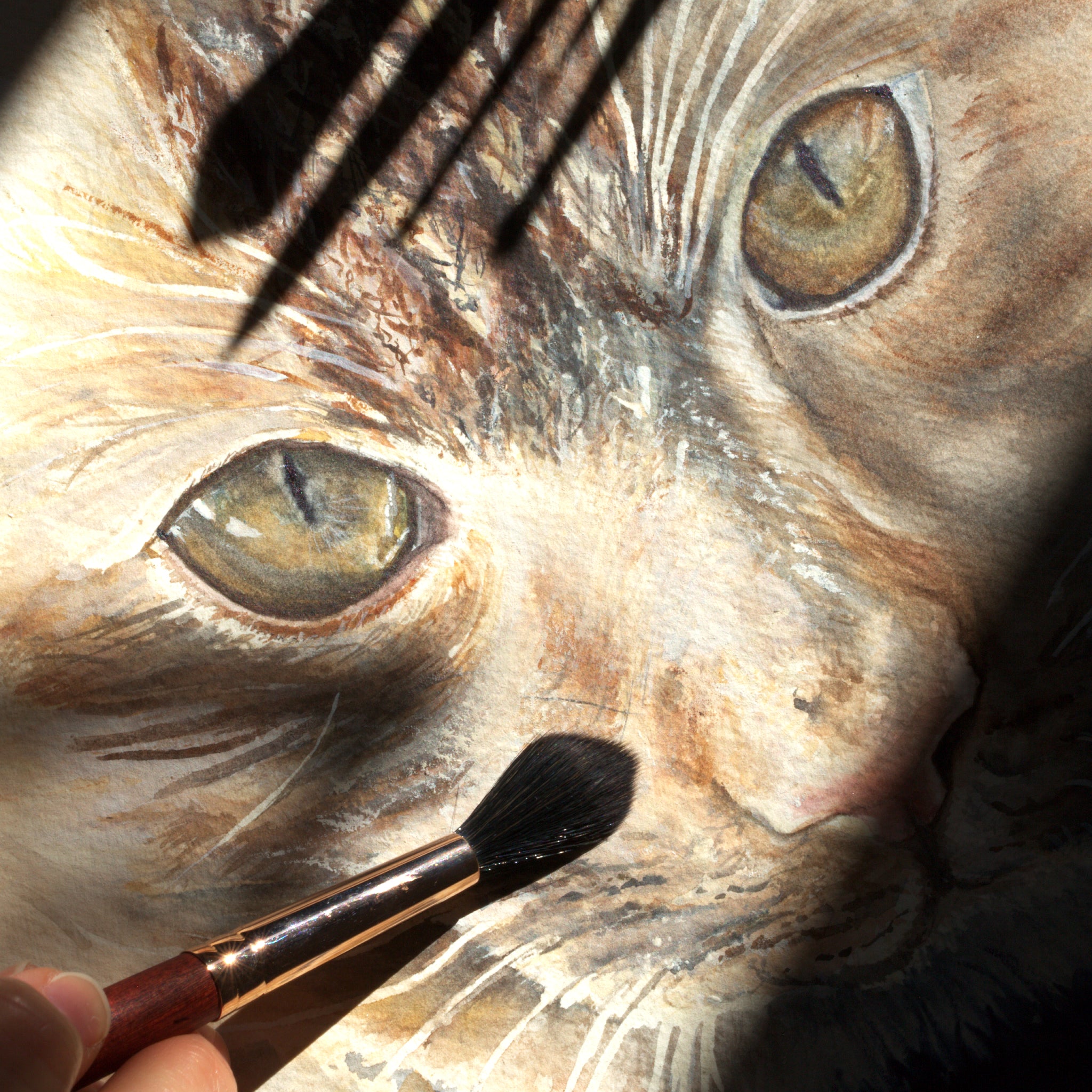 Watercolor tabby cat close-up with burgundy paintbrush adding facial details to the painting.