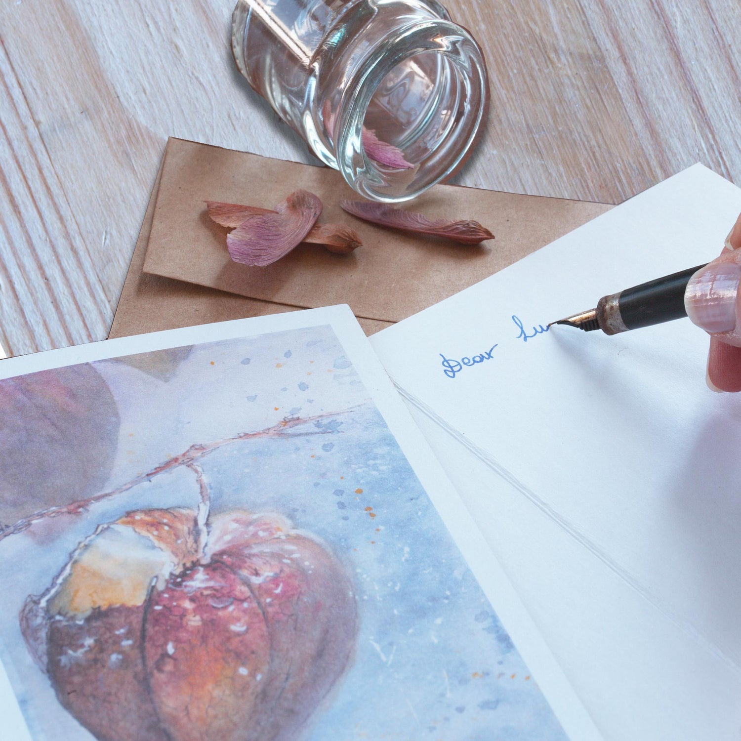 Close-up of a Chinese lantern watercolor greeting card on top of an open card, with a hand holding a fountain pen writing on it. Envelope peeks out from underneath, and dried seed pods are casually fanned out from a small glass jar on a wooden surface.