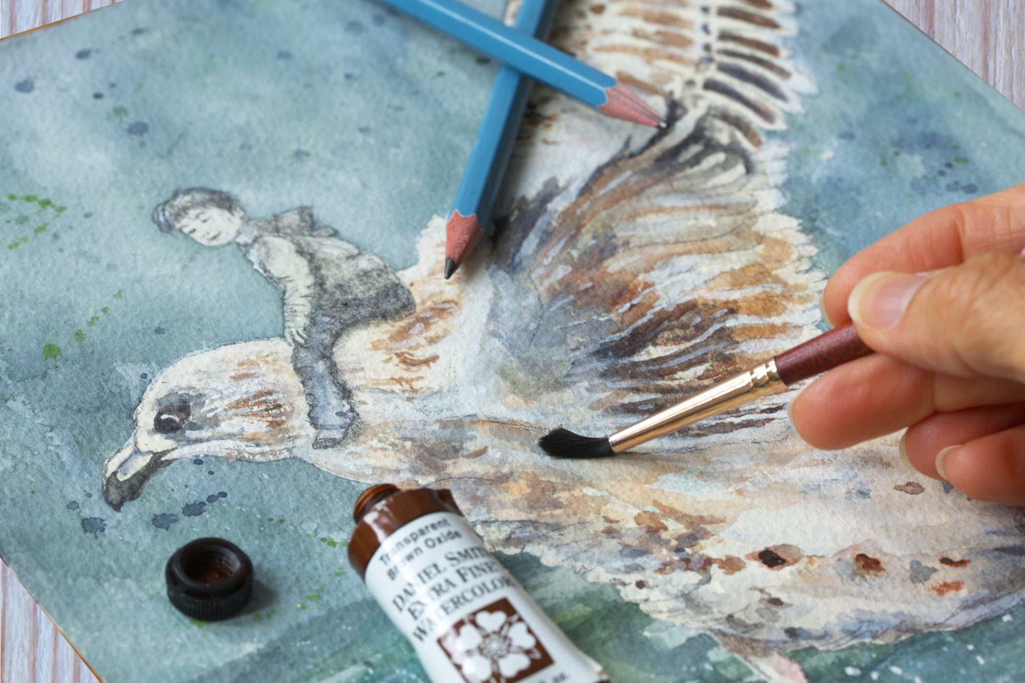 Closeup of work-in-progress watercolor painting of a person riding a seagull on textured paper, with hands painting it and brushes and a watercolor tube on the painting.