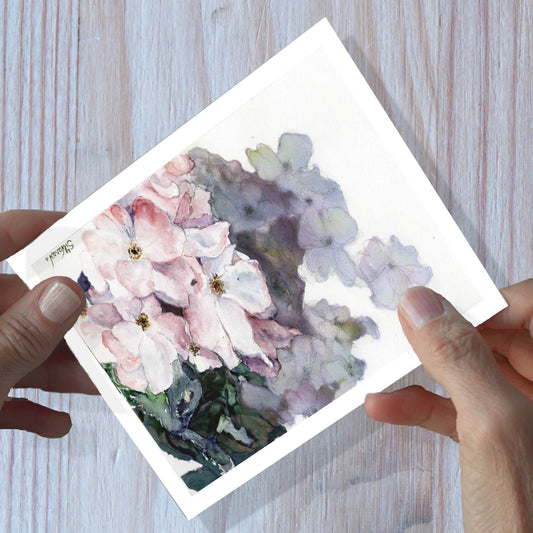 Hands holding a wild rose watercolor greeting card, shown on a wooden surface.