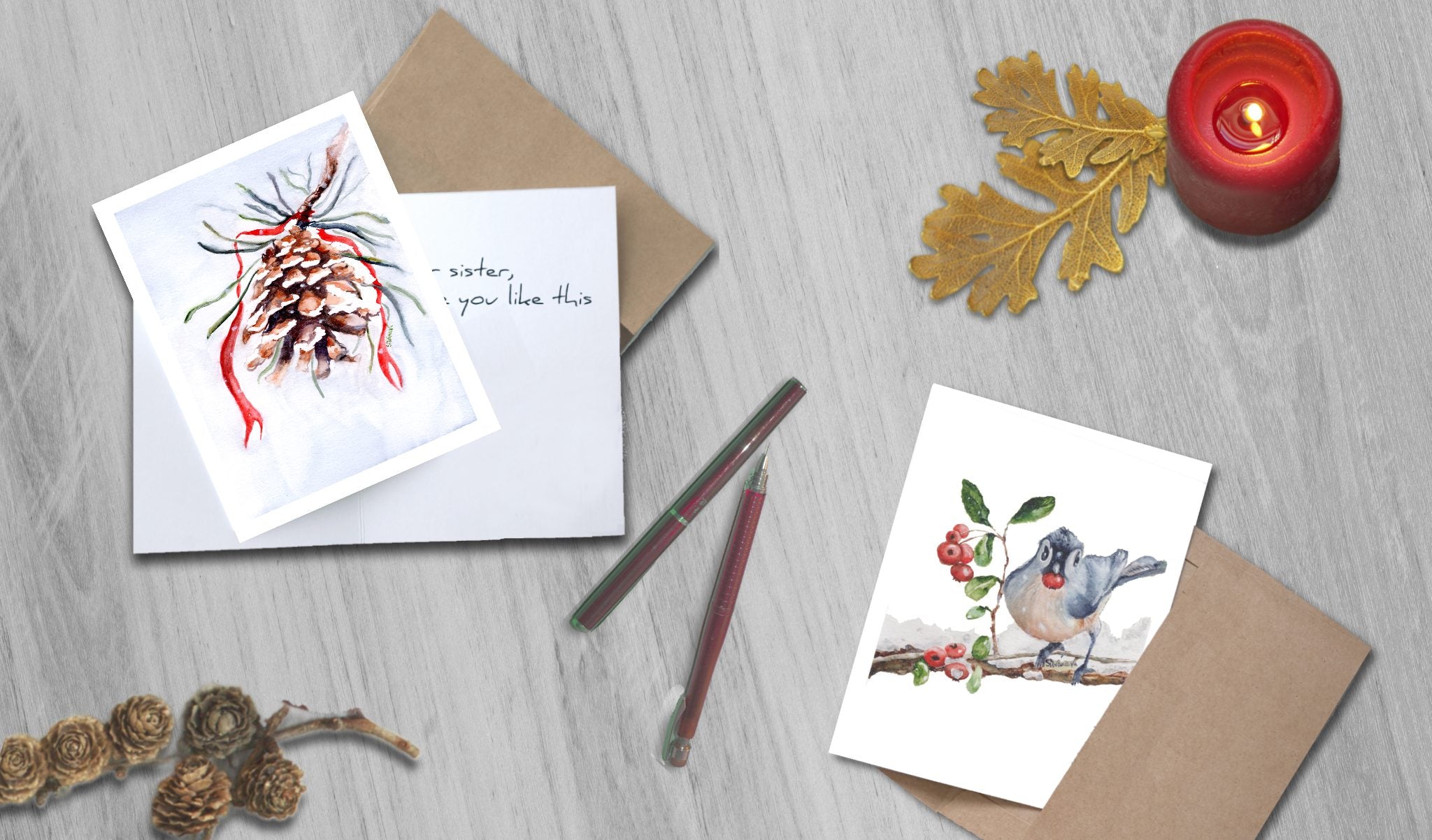Two watercolor print greeting cards with a pinecone and a bluejay bird  designs on a wooden surface with a candle and decorative elements.
